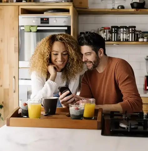 Image of a man and a woman leaning against their kitchen counter in front of their breakfast while looking at the cell phone the man is holding, both smiling