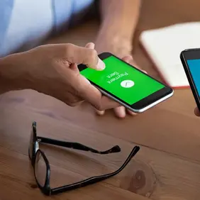 Two people hold smartphones showing payment sent and money received confirmations over a wooden table with a notebook and glasses nearby.