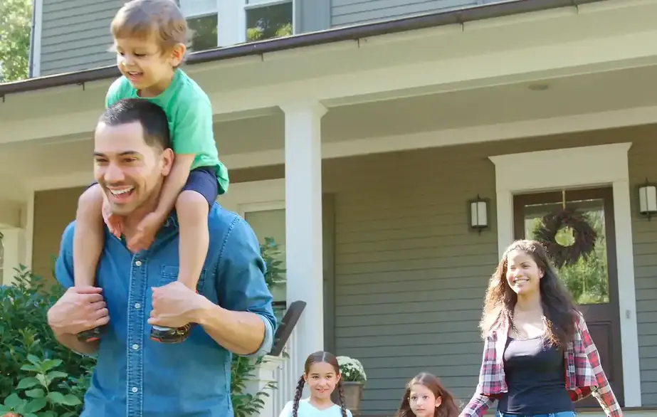 Image of a happy family walking away from the front of their house, the man in the foreground with a child on his shoulders and the woman and two little girls following a few feet behind 