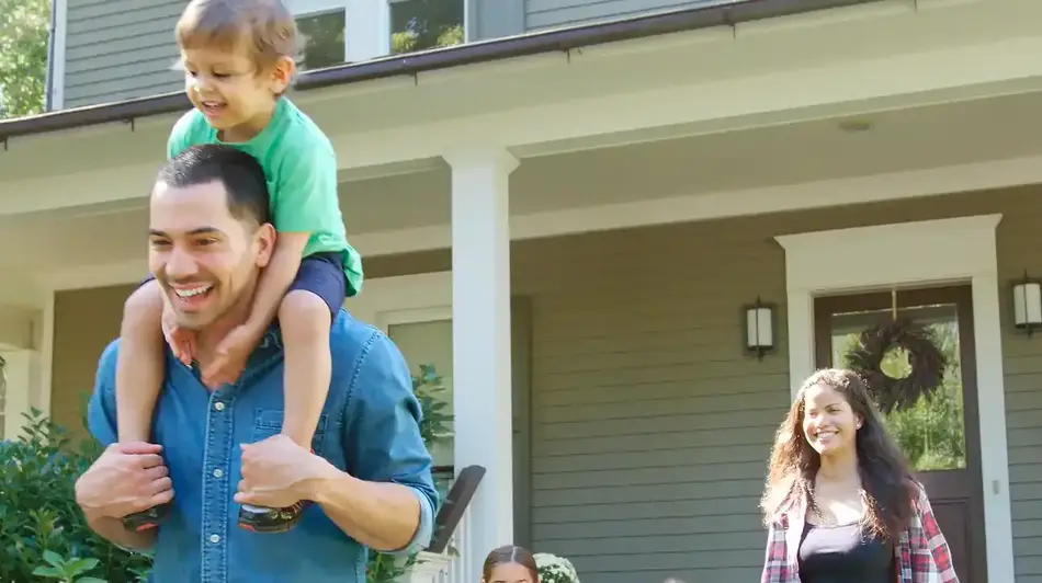 Image of a happy family walking away from the front of their house, the man in the foreground with a child on his shoulders and the woman and two little girls following a few feet behind 