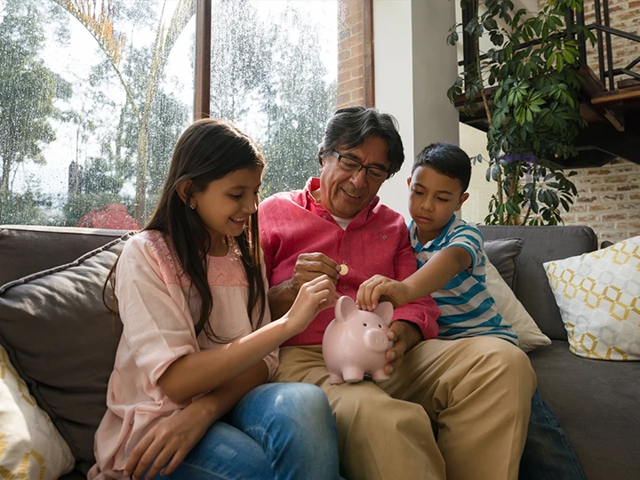 Image of a father sitting on a couch between his daughter and son while they add coins to a piggy bank sitting on dad's leg