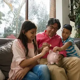 Image of a father sitting on a couch between his daughter and son while they add coins to a piggy bank sitting on dad's leg