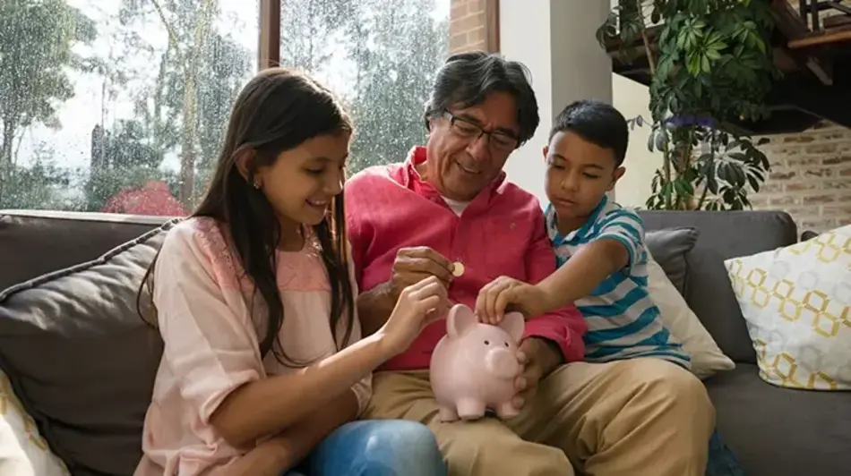Image of a father sitting on a couch between his daughter and son while they add coins to a piggy bank sitting on dad's leg