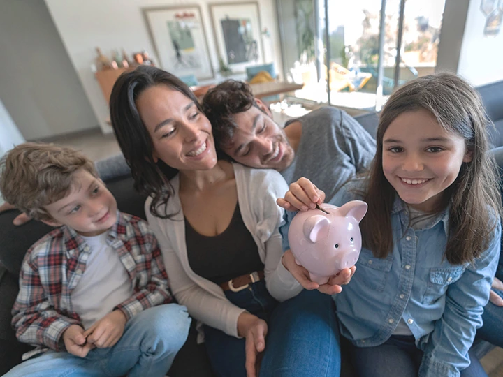 Image of a man and a woman with their two children. Mom is holding a piggy bank while her smiling daughter is dropping a coin into the slot.