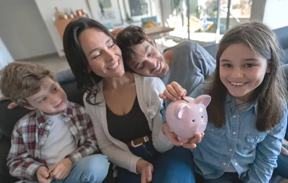 Image of a man and a woman with their two children. Mom is holding a piggy bank while her smiling daughter is dropping a coin into the slot.