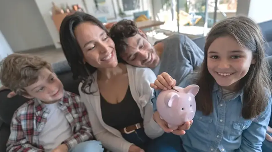 Image of a man and a woman with their two children. Mom is holding a piggy bank while her smiling daughter is dropping a coin into the slot.
