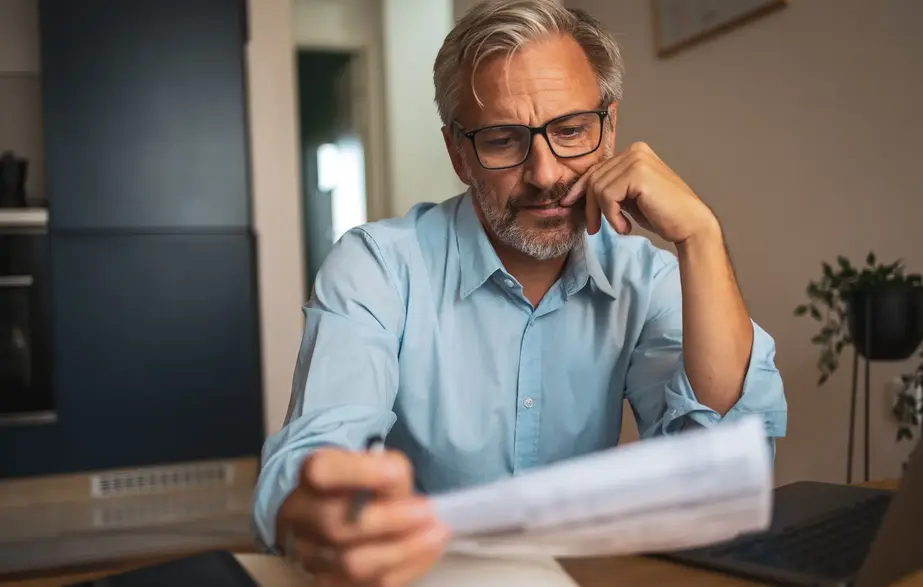 Gray-haired man wearing glasses reviews a document at a table next to a laptop, looking thoughtful in a home setting.