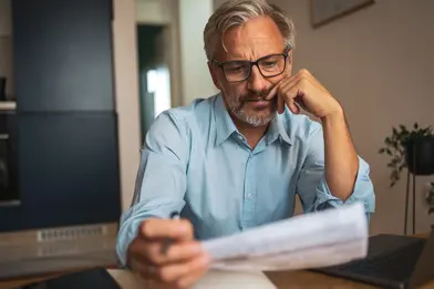 Gray-haired man wearing glasses reviews a document at a table next to a laptop, looking thoughtful in a home setting.