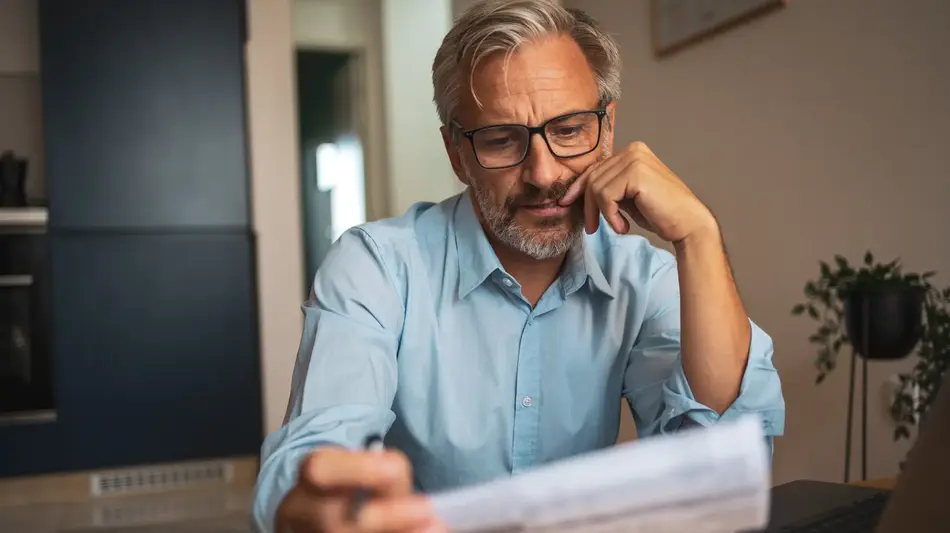 Gray-haired man wearing glasses reviews a document at a table next to a laptop, looking thoughtful in a home setting.