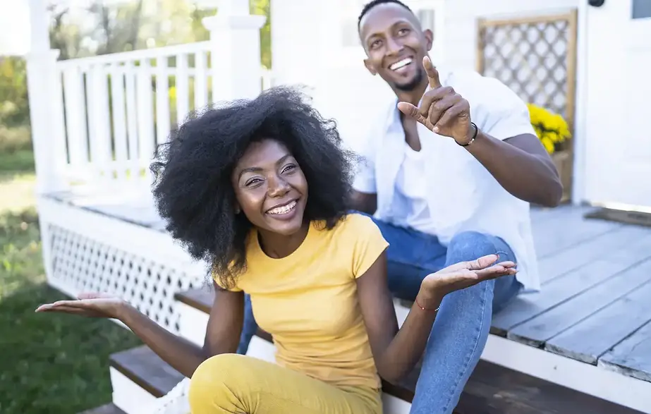 A man and a woman are smiling, sitting on the steps of their front porch, with the man pointing toward the camera