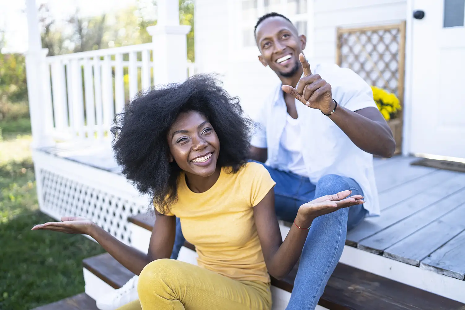 A man and a woman are smiling, sitting on the steps of their front porch, with the man pointing toward the camera