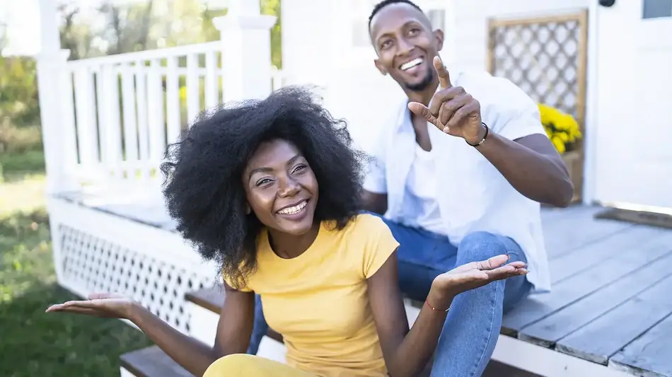 A man and a woman are smiling, sitting on the steps of their front porch, with the man pointing toward the camera