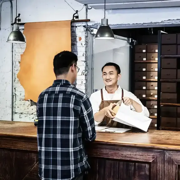 A shop owner chats with a customer from across the counter while packaging the item he bought