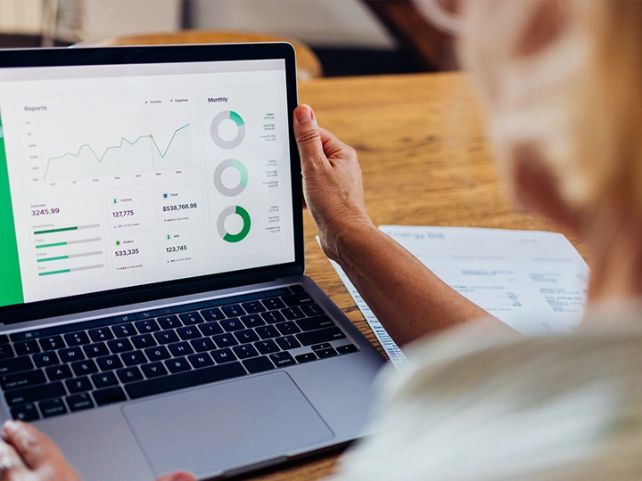 Image looking from behind an older woman's left shoulder toward her open laptop with her investments dashboard showing