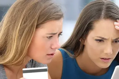Two women looking worried while looking at a laptop computer screen, with one of the women holding a credit card.
