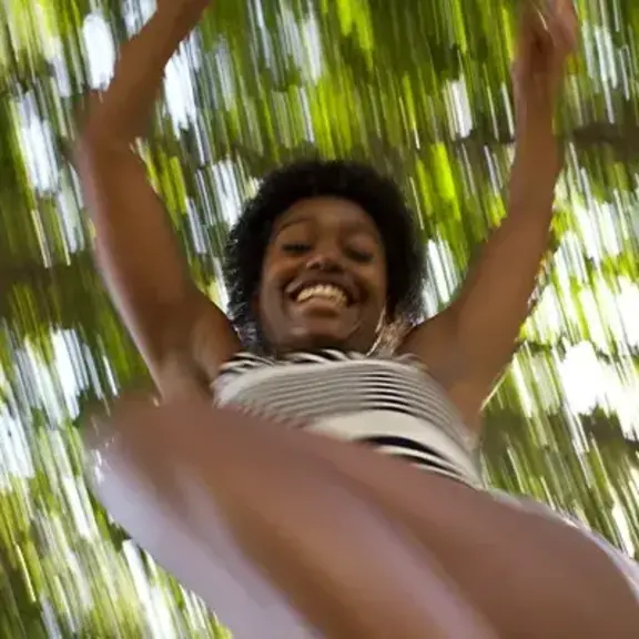 Smiling woman outdoors, viewed from below, with arms raised against a background of green trees and sunlight.