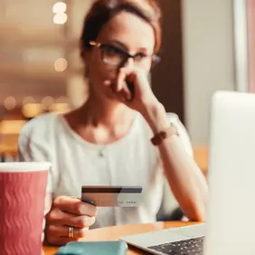 Woman wearing glasses holds a credit card while looking at a laptop at a café table, with a coffee cup nearby.