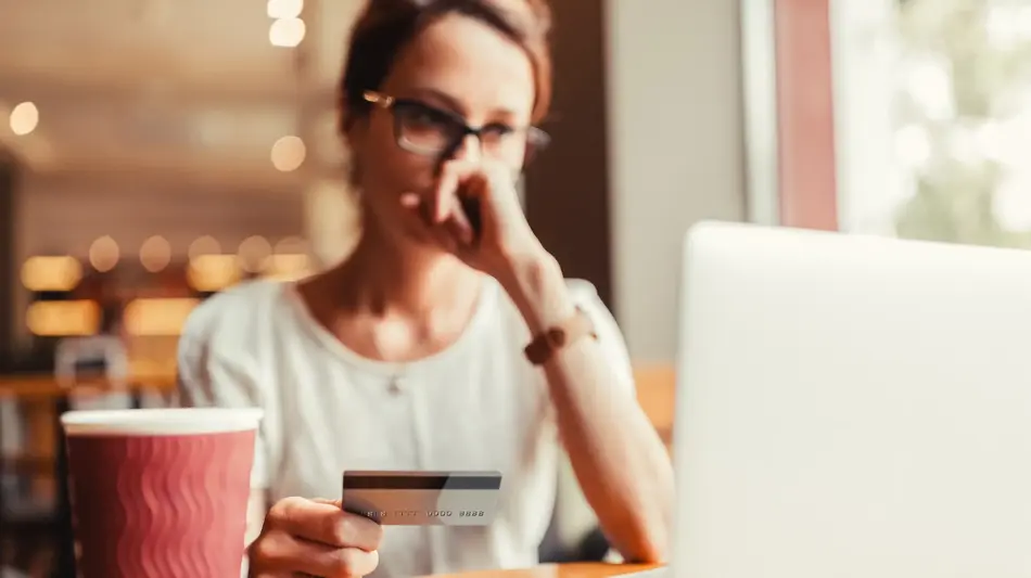 Woman wearing glasses holds a credit card while looking at a laptop at a café table, with a coffee cup nearby.