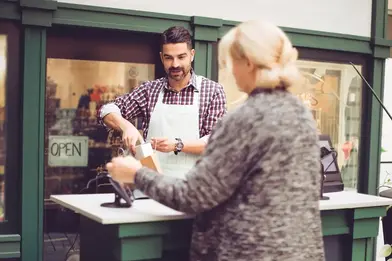 A woman swipes her Everwise card at an outside store counter while the shop owner packages the small item she bought