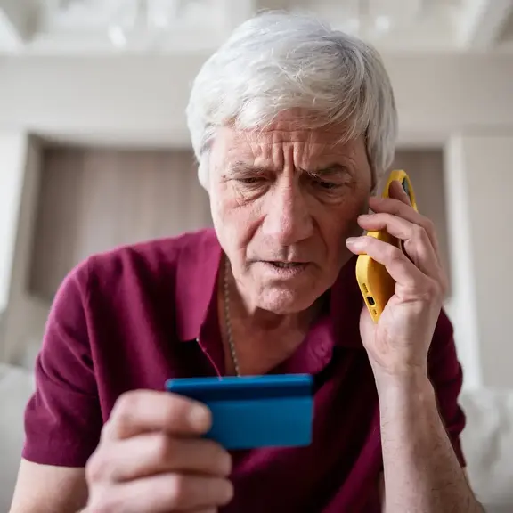 Image of an older man with a concerned look on his face while looking at his credit card while talking on the phone.