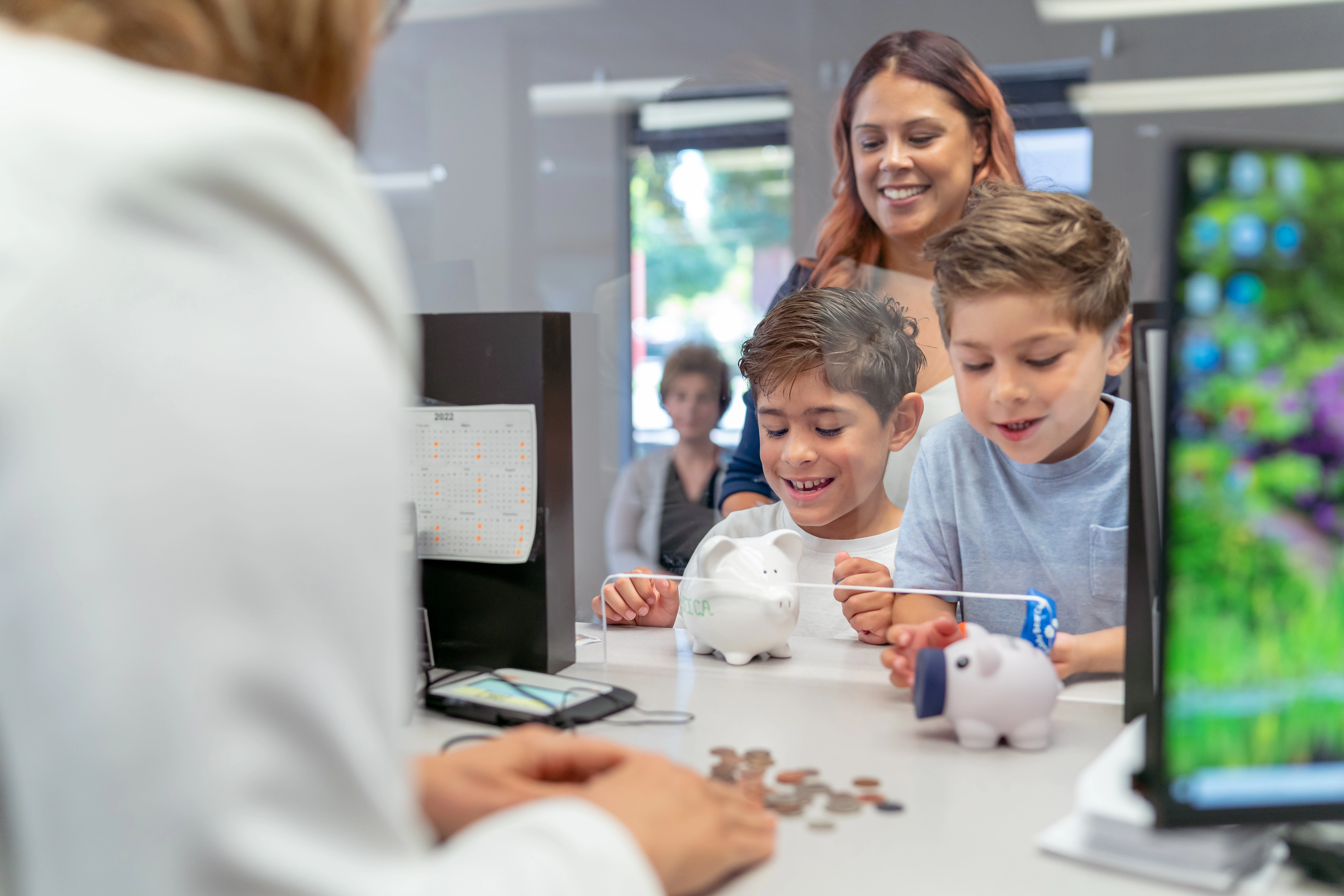 Two elementary age boys of Hispanic descent smile with excitement as a bank teller counts the coins they brought from their piggy banks.