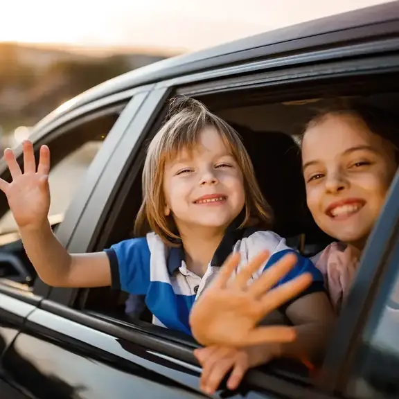 Two children sticking their heads out of the car window and waving at the camera