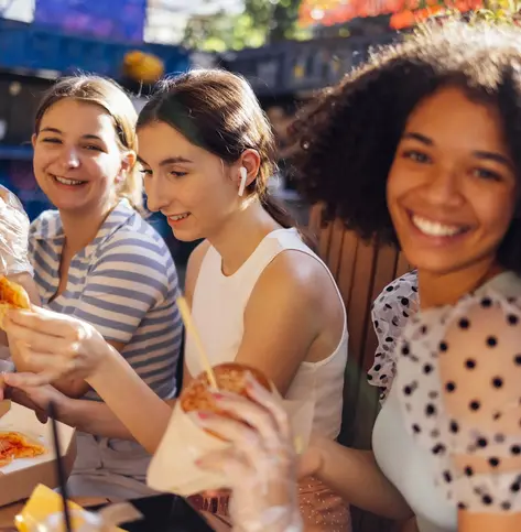 Cute smiling teenage girls are sitting in open air cafe and eating fast food