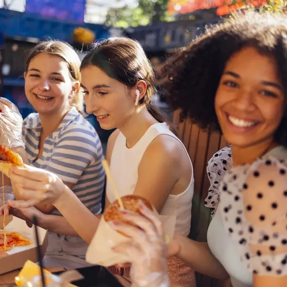 Cute smiling teenage girls are sitting in open air cafe and eating fast food