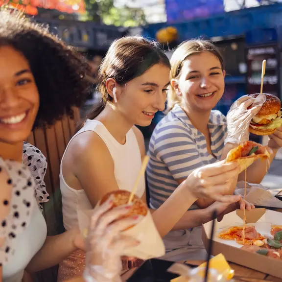 Cute smiling teenage girls are sitting in open air cafe and eating fast food