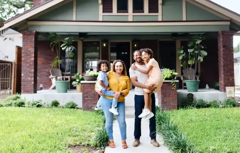 A man and woman smiling, standing in front of their new home, each holding a child in their arms