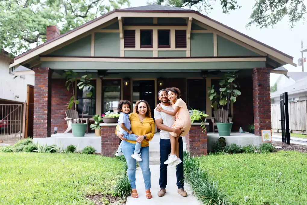 A man and woman smiling, standing in front of their new home, each holding a child in their arms