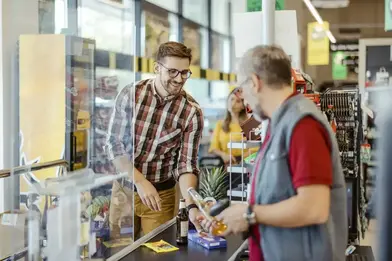 A smiling customer places items on the checkout belt while the cashier checks the price of an item